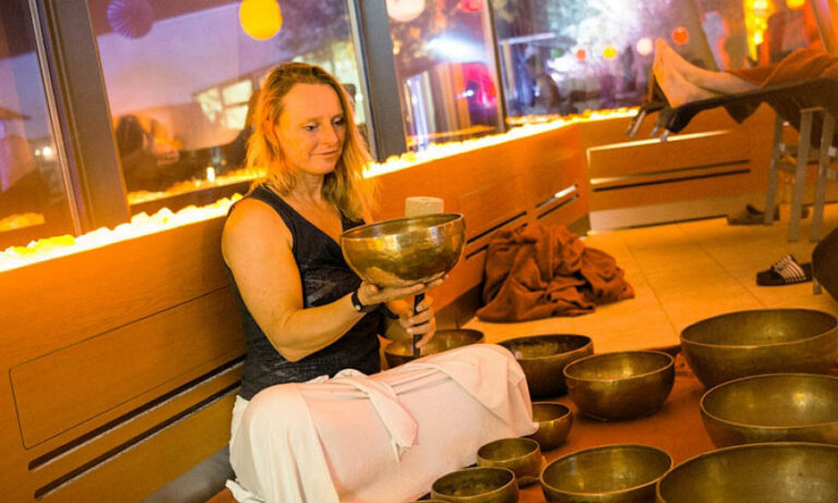 Frau mit KLangschalen sitzt im Schneidersitz vor eine Gruppe Teilnehmer